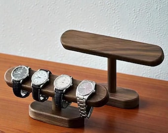 May include: Wooden watch display stand with four wristwatches. The watches have black leather and silver metal bands. The stand is made of dark brown wood and has a minimalist design.