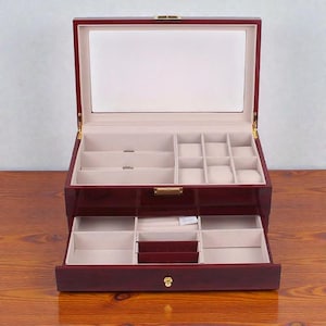 May include: A cherry-red wooden jewelry box with a glass top and multiple compartments. The interior is lined with a cream-colored fabric, and the box has a pull-out drawer. The box is open, revealing its storage spaces.