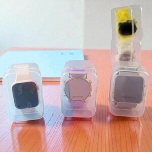 Pode incluir: Três relógios LED em caixas de plástico transparente. Um relógio tem um mostrador quadrado preto e uma pulseira rosa claro. Outro tem um mostrador redondo rosa com o texto "LED WATCH" e uma pulseira roxa. O terceiro relógio tem um mostrador quadrado cinza.