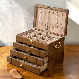 May include: A wooden jewelry box with multiple drawers and compartments. The box is open, revealing necklaces hanging from the lid and various jewelry pieces in the compartments. The box has a gold-colored handle and drawer pulls.