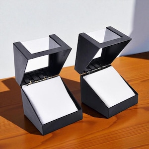 May include: Two black, open-top display boxes with white interiors sit on a wooden surface. The boxes are angled, with a hinged lid and a square opening at the top. The interior of each box is lined with white fabric.