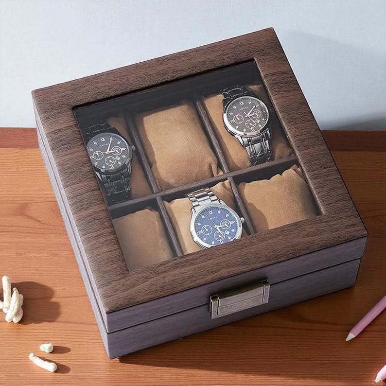 May include: A wooden watch display case with a glass top, showcasing four wristwatches. The case has a dark brown finish with six compartments, each containing a tan pillow to hold the watches. The watches have black and silver bands and blue and black faces.