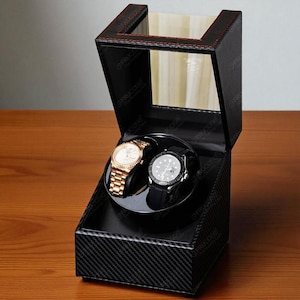 May include: Black watch winder with a carbon fibre pattern, holding two wristwatches. One watch has a gold band, the other a black band. The winder has a clear top and red stitching.