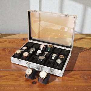 Pode incluir: Uma mala de exibição de relógios prateada com tampa transparente, aberta para revelar uma coleção de vários relógios de pulso. A mala é forrada com veludo preto e tem compartimentos individuais para cada relógio. Dois relógios repousam em suportes de veludo preto em frente à mala.