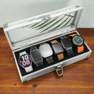 Pode incluir: Uma caixa de exibição de relógios prateada com seis smartwatches. Os relógios têm pulseiras de várias cores, incluindo branco, preto, castanho e laranja. Os mostradores são pretos, prateados e verdes. A caixa tem uma tampa transparente e um interior preto.