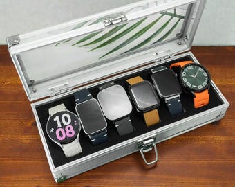 May include: A silver watch display case with six smartwatches. The watches have various band colors, including white, black, brown, and orange. The watch faces are black, silver, and green. The case has a clear lid and a black interior.