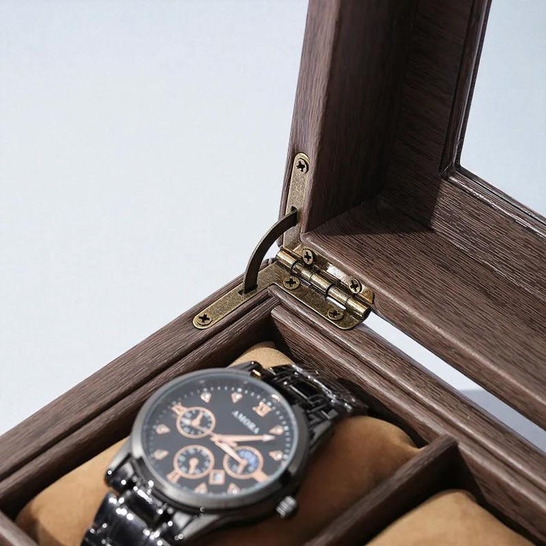 May include: A wooden watch display case with a hinged lid and brass hardware. Inside, a black watch with a black metal band rests on a tan cushion. The watch face has gold accents and the brand name "AMORA".