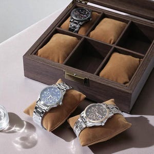 May include: A wooden watch box with six compartments, each containing a brown pillow. Three silver watches with various face designs are displayed. One watch is inside the box, and two are on pillows outside the box.