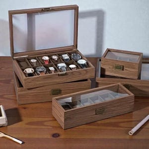 May include: Wooden watch display boxes with clear glass tops. The open box reveals a collection of watches. Additional closed boxes are visible. The boxes are made of wood with a light brown finish and brass-colored hardware.
