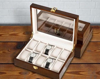 May include: A wooden watch box with a glass top, showcasing several wristwatches with black leather bands. The interior features cream-colored cushions to hold the watches. The box is open, revealing the watches, and sits on a wooden surface.