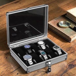 Könnte beinhalten: Geöffnete silberne Uhrenbox mit transparentem Deckel, die sechs Armbanduhren mit verschiedenen Designs zeigt. Der Innenraum ist mit schwarzem Samt ausgekleidet. Die Box steht auf einer Holzoberfläche, mit einem Buch und einer Lupe im Hintergrund.