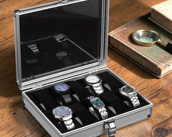 May include: A silver watch case with a clear lid, open to reveal six wristwatches with various band and face designs. The interior is lined with black velvet. The case sits on a wooden surface, with a book and magnifying glass in the background.