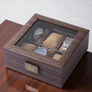 May include: A wooden watch box with a glass top, showcasing four wristwatches. The box is a dark brown color with a rectangular shape and a metal clasp. The interior features soft, tan-colored cushions to hold the watches.
