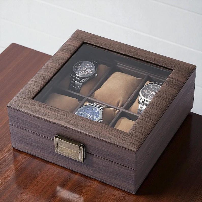 May include: A wooden watch box with a glass top, showcasing four wristwatches. The box is a dark brown color with a rectangular shape and a metal clasp. The interior features soft, tan-colored cushions to hold the watches.