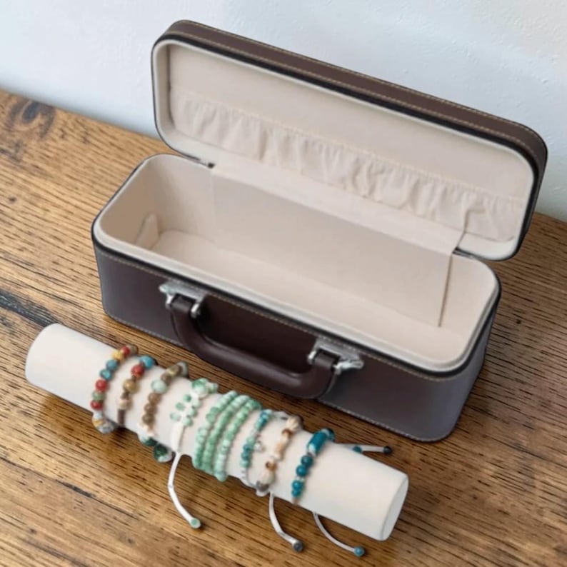 Puede incluir: Un estuche de joyer&iacute;a de cuero marr&oacute;n abierto con un interior crema descansa sobre una superficie de madera. Una pantalla cil&iacute;ndrica blanca contiene varias pulseras de cuentas en varios colores, incluyendo verde, azul y rojo.