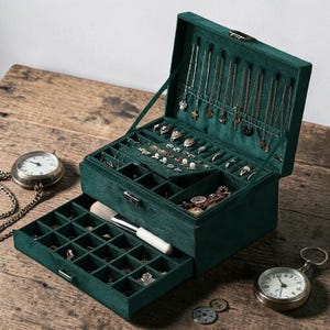 May include: A dark green velvet jewelry box with multiple compartments and a pull-out drawer. The box is open, displaying various necklaces, rings, and other jewelry. A makeup brush rests in the drawer. Two pocket watches and coins are on the wooden surface.