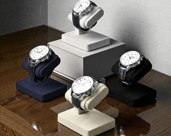 May include: Five silver-toned watches with black leather bands are displayed on individual stands. The watch faces are white with black markings. The stands are in various colors, including black, navy blue, and cream.
