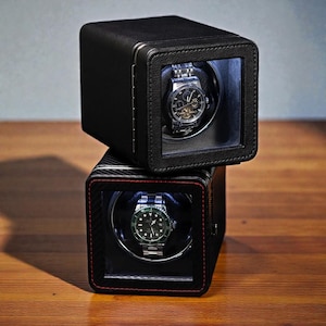 May include: Two black watch winders stacked on top of each other. Each winder has a clear window displaying a luxury wristwatch. The top winder has a leather texture, while the bottom one has a carbon fiber design with red stitching.