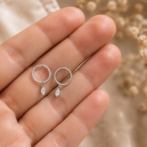 May include: A pair of silver hoop earrings, each featuring a small, dangling marquise-cut clear gemstone charm. The earrings are crafted from a polished silver-toned metal, showcasing a simple circular hoop design with a delicate, faceted stone.