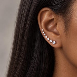 May include: A silver-toned ear climber earring featuring seven round, clear, faceted stones arranged in a graduated size sequence that follows the natural curve of the earlobe, secured with a metal post and backing mechanism.