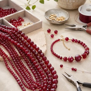 May include: A collection of faceted deep red gemstone beads arranged in long strands alongside a finished bracelet with gold-toned metal spacers and a clasp. Jewelry making tools including pliers and loose beads are present in a divided tray.