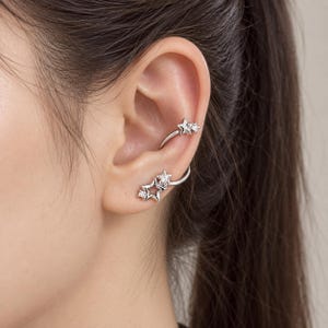May include: A silver-toned ear cuff and earring set featuring star-shaped designs. The upper ear cuff displays a small star with a clear stone, while the lower earring showcases a larger open star outline paired with a smaller star and clear stone.