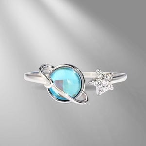 May include: A silver ring with a blue, planet-shaped central stone, encircled by a band. A star, set with small, clear stones, sits on one side. The ring has an open design.