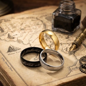 May include: Three metal rings in gold, silver, and black tones, each featuring an engraved script pattern around the outer band. The rings are arranged together on a vintage-style map that includes the text Minas Tirith.