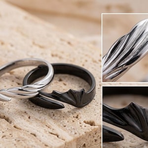 May include: A pair of open-ended rings, one in polished silver-tone metal and one in black metal. Each ring features a textured, wing-like design that tapers to a point. The silver ring has a feathered texture, while the black ring has a smooth finish.