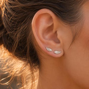 May include: Two silver-toned stud earrings shaped like feathered wings, each featuring small embedded clear stones along the surface. The earrings are positioned horizontally on the earlobe, with one wing pointing slightly upward and the other downward.