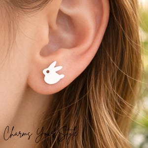 May include: A silver bunny-shaped stud earring is worn on an ear. The earring is a simple, minimalist design with a small, round hole representing the eye. The text "Charms Your Style" is at the bottom left.