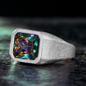 May include: A silver ring featuring a large, square-cut, multi-coloured gemstone. The gemstone displays a spectrum of colours, including green, blue, purple, and yellow. The ring has a textured band and a modern, geometric design.