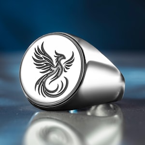 Può includere: Un anello con sigillo d'argento con un design dettagliato di fenice. L'anello presenta una fascia lucida e arrotondata e una faccia circolare con una fenice incisa in nero. La fenice è raffigurata con le ali spiegate e una coda curva, su sfondo bianco.