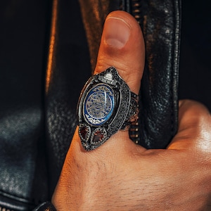 Peut inclure: Une bague en argent détaillée avec une pierre centrale ovale bleue ornée d'un motif doré. Deux pierres rouges flanquent le centre bleu. La bague présente des motifs complexes et est portée sur un pouce.
