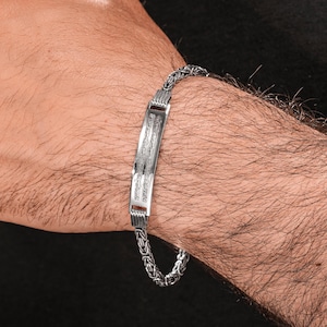May include: A silver bracelet with a rectangular center plate and a chain-link band. The center plate has engraved text. The bracelet is worn on a wrist.