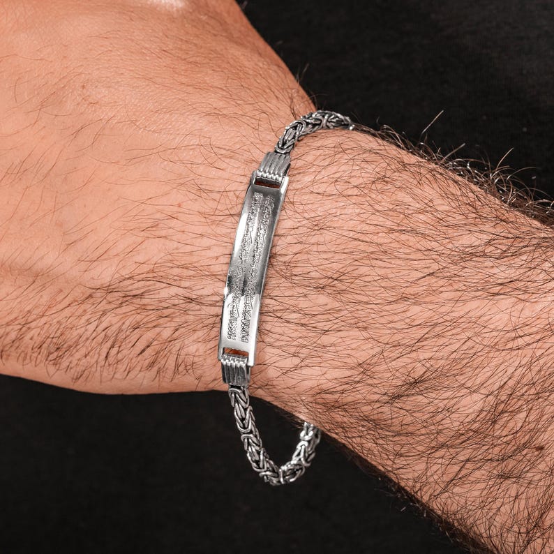 May include: A silver bracelet with a rectangular center plate and a chain-link band. The center plate has engraved text. The bracelet is worn on a wrist.
