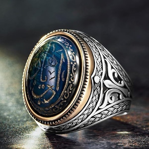 May include: A silver ring with an oval blue gemstone. The gemstone has gold-colored calligraphy. The ring has a gold-colored band around the gemstone and intricate silver detailing on the band.