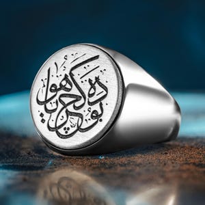 May include: A silver signet ring with an engraved Arabic inscription. The ring has a polished, rounded band and a circular face with the black text. The background is a gradient of blue and brown.