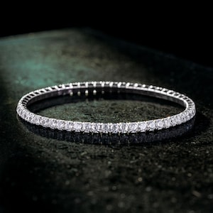 May include: A silver bracelet with a continuous row of sparkling, clear gemstones. The bracelet is set against a dark, reflective surface, highlighting the brilliance of the stones. The jewelry piece is a classic design.