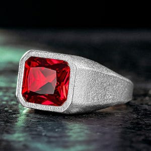 May include: A silver ring with a large, square, deep red gemstone. The ring has a textured band and a bezel setting that holds the gemstone securely. The gemstone is faceted, reflecting light.