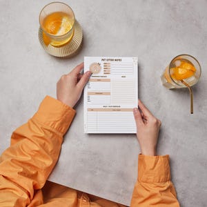 May include: A pet sitter notes notepad with the title "PET SITTER NOTES" is held by a person wearing an orange shirt. The notepad has sections for contact information, daily schedule, and notes. Two glasses of orange-infused water are on the table.