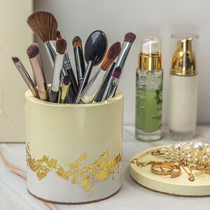 May include: A cylindrical makeup brush holder with a cream and gold leaf design, filled with various makeup brushes. Beside it are two cosmetic bottles, a small tray with jewelry, and a pearl necklace.
