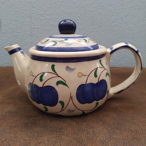 May include: A white ceramic teapot with a blue and green floral design featuring blue plums. The teapot has a lid with a blue knob.
