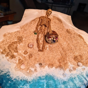 May include: A skeleton figurine holding a treasure chest with jewels, standing on a beach scene with sand and blue ocean waves.