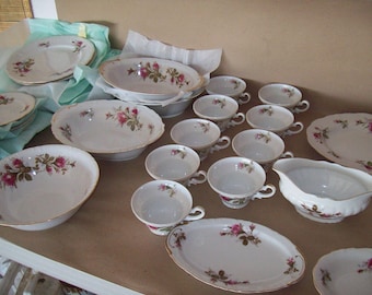 Może przedstawiać: Zestaw białej porcelanowej zastawy stołowej z kwiatowym wzorem z różowymi różami. Zestaw zawiera talerze, miski i sosjerkę.