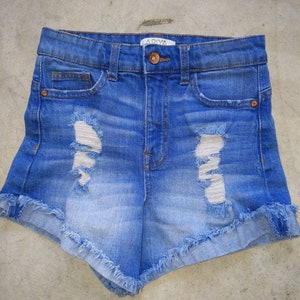May include: Light blue denim shorts with frayed edges and distressed details. The shorts have a high waist and a button and zipper closure. The label on the shorts reads "ARIYA".