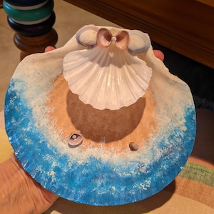 May include: A blue and white seashell dish with a smaller seashell on top. The dish is painted to look like an ocean wave crashing on a beach.