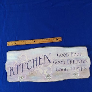 May include: A wooden sign with a purple and white background. The sign says "Kitchen" in purple lettering and "Good Food, Good Friends, Good Times" in black lettering. The sign has three decorative knobs.