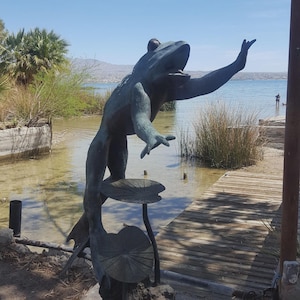 May include: A bronze frog statue with one arm raised, standing on a lily pad. The statue is near a body of water with a wooden dock and vegetation in the background. The frog's mouth is open, and it appears to be singing.