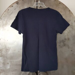 Puede incluir: Una camiseta negra lisa de cuello en V colgada de una percha. La camiseta est&aacute; hecha de una tela suave y c&oacute;moda.